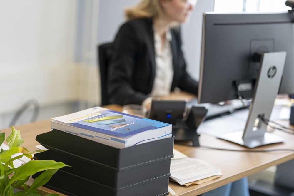 Een stapel documentenbakjes met daarop de 'Smartengeld' gids op een bureau, terwijl een medewerker in de onscherpe achtergrond achter een computer werkt.