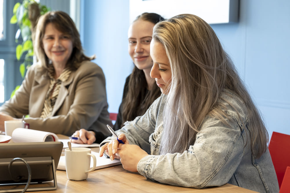 Een vrouw zit aandachtig aan een houten tafel met een kop koffie, terwijl twee glimlachende vrouwelijke collega's op de achtergrond toekijken tijdens een overleg.