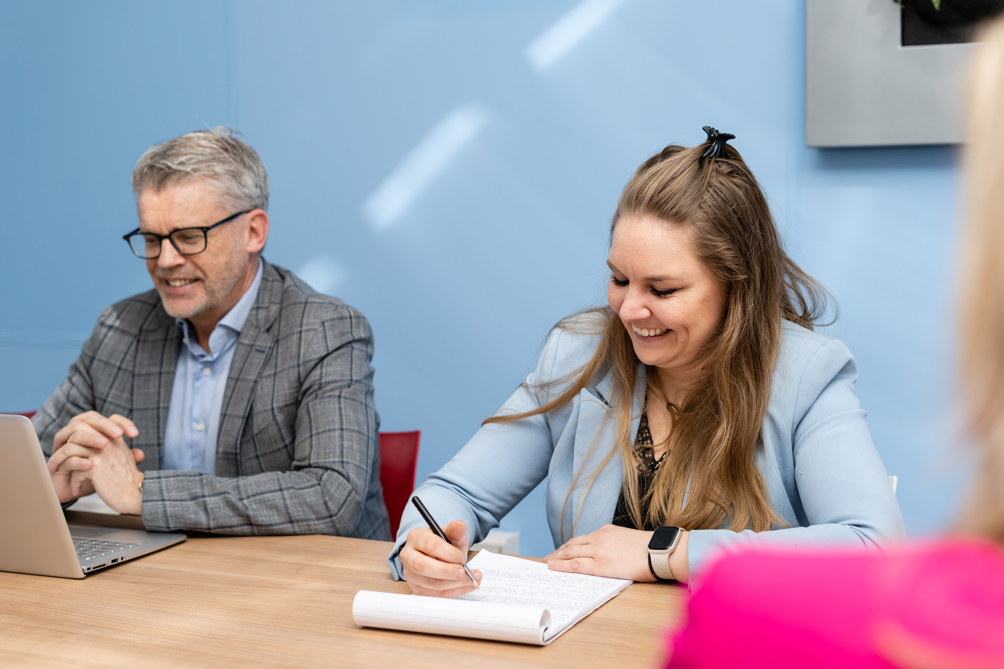 Een lachende vrouw maakt aantekeningen tijdens een gesprek, terwijl een man naast haar zit aan een houten tafel.