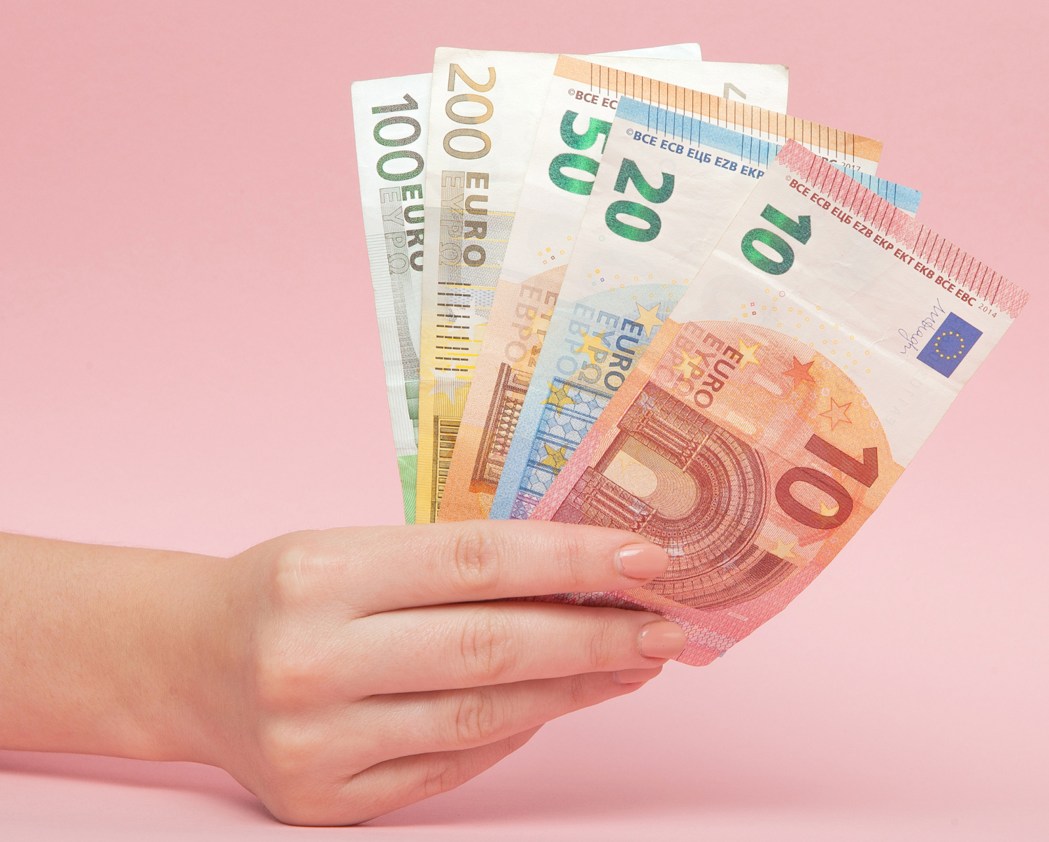 Een hand houdt een waaier van diverse eurobiljetten (10, 20, 50, 100 en 200 euro) vast tegen een effen roze achtergrond.