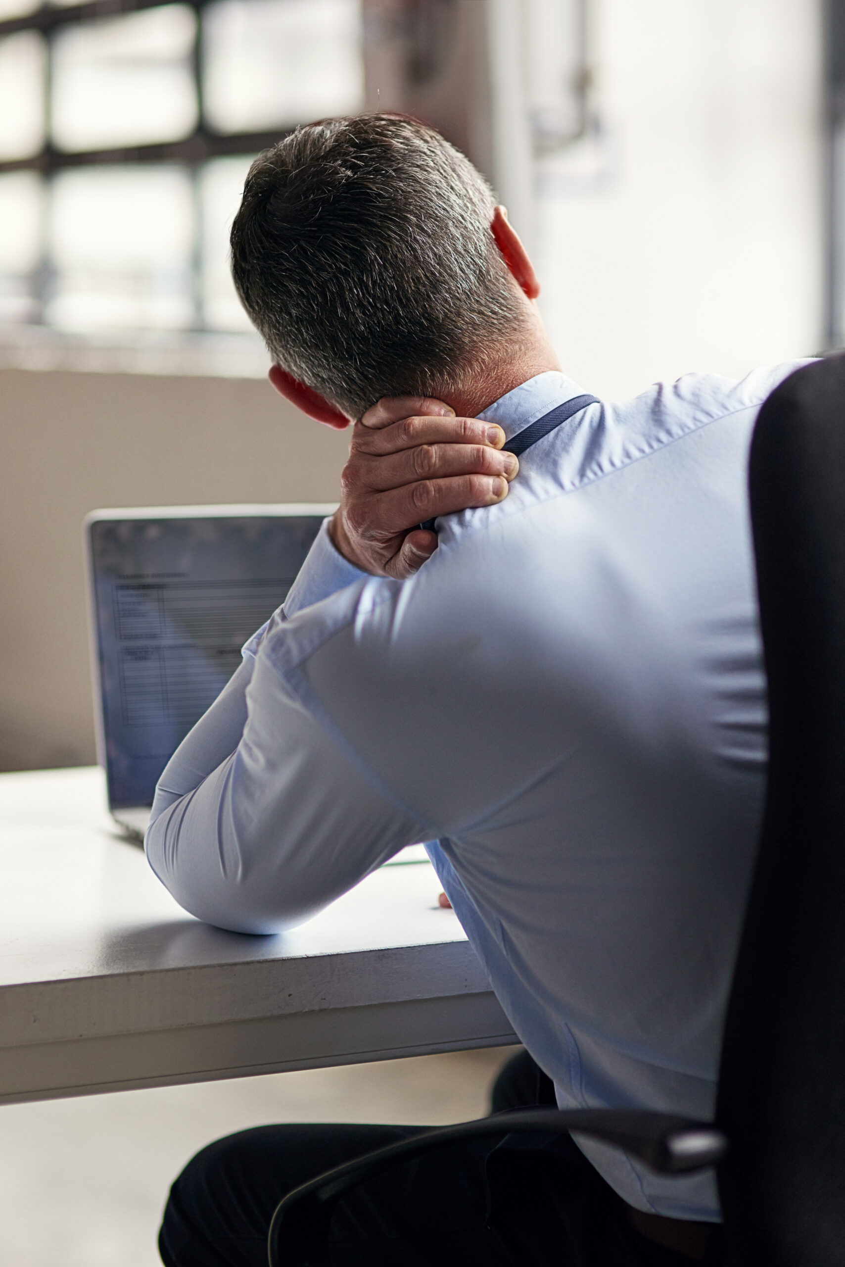 Man zit achter een bureau met een laptop en houdt zijn nek vast, wat wijst op nek- of spierpijn tijdens het werken.