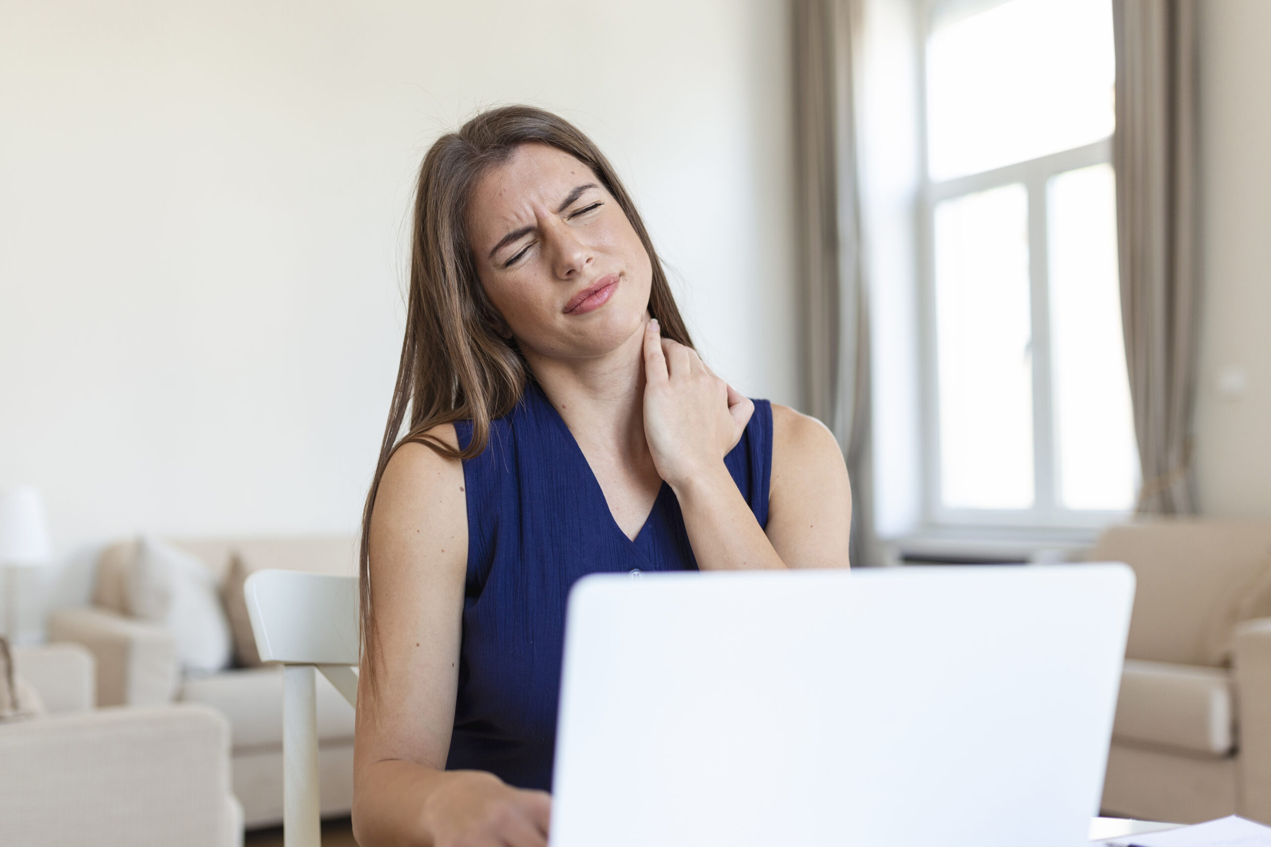 Vrouw zit achter een laptop en houdt haar nek vast, met een pijnlijke gezichtsuitdrukking, alsof ze last heeft van nekklachten tijdens het werken.
